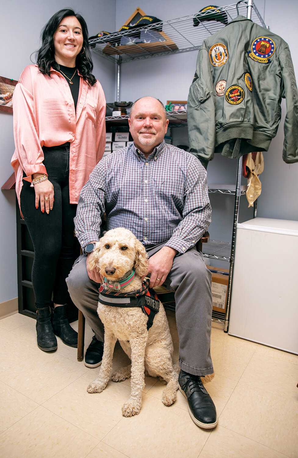 Two members of the FICOM team with a service dog, beside a flight jacket and military memorabilia
